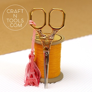 May include: Gold-coloured scissors with a pink Eiffel Tower tassel. The scissors are sitting on a spool of yellow thread.