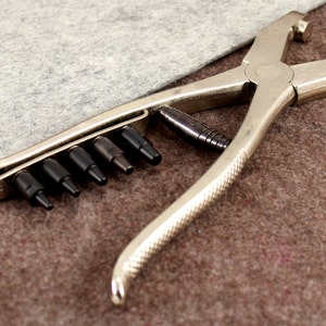 May include: A silver metal punch tool with a handle and a set of interchangeable punches. The tool is designed for creating holes in leather or fabric.