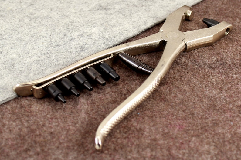May include: A silver metal punch tool with a handle and a set of interchangeable punches. The tool is designed for creating holes in leather or fabric.