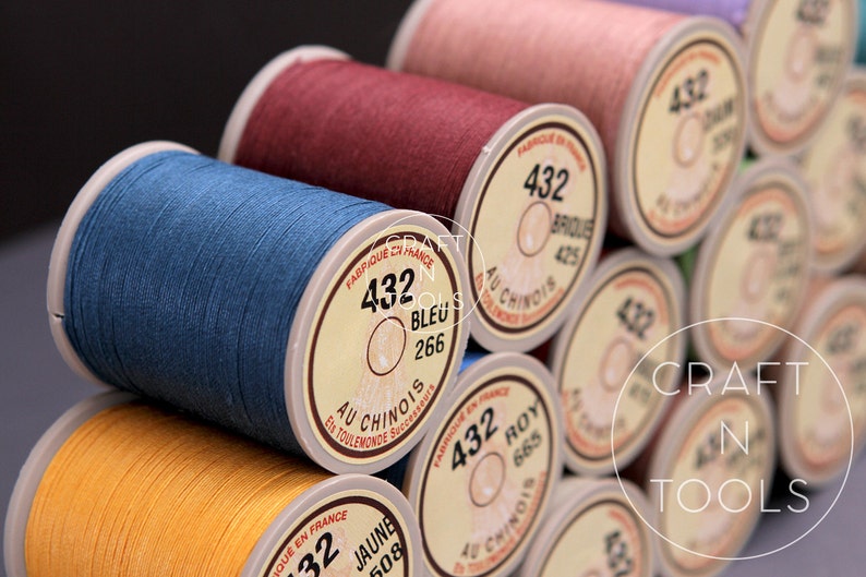 May include: Close-up of spools of thread in various colors, including blue, red, yellow, and brown. Each spool has a label with the number "432" and the color name in French. The label also includes the text "Au Chinois" and "Fabrique en France".
