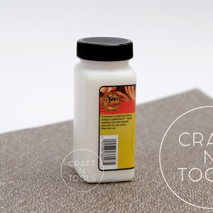 K&ouml;nnte beinhalten: Eine wei&szlig;e Flasche mit schwarzem Deckel, die Fiebing's Lederfarbe enth&auml;lt. Die Flasche ist mit dem Produktnamen und Gebrauchsanweisungen beschriftet. Die Flasche steht auf einer braunen, strukturierten Oberfl&auml;che.