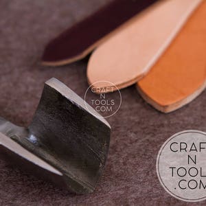 Puede incluir: Primer plano de una herramienta de trabajo de cuero de metal con un borde afilado. La herramienta est&aacute; sobre una superficie marr&oacute;n con tres piezas de cuero en el fondo. El cuero est&aacute; en tonos de marr&oacute;n, melocot&oacute;n y burdeos. El texto "CRAFT N TOOLS .COM" es visible en la superficie.