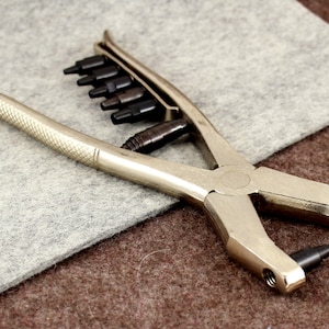 May include: A silver metal punch tool with a set of interchangeable punches. The tool has a handle and a lever to operate the punches. The punches are arranged in a row on the tool.