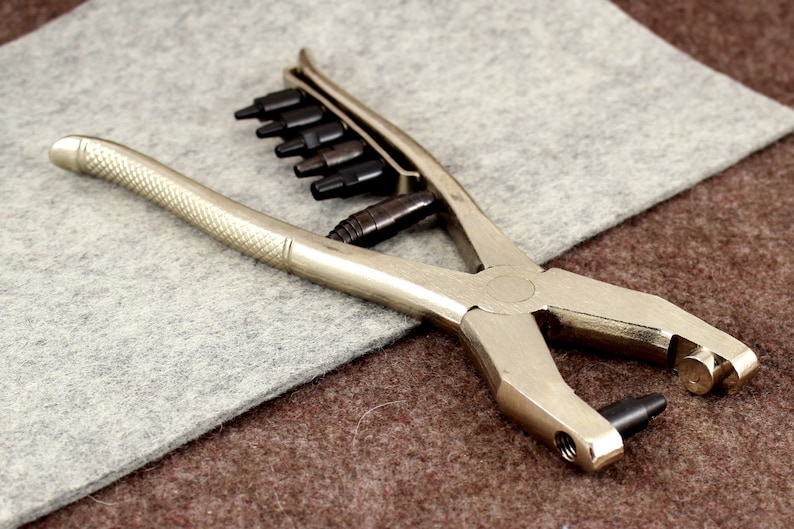 May include: A silver metal punch tool with a set of interchangeable punches. The tool has a handle and a lever to operate the punches. The punches are arranged in a row on the tool.