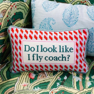 May include: A rectangular decorative pillow with the text "Do I look like I fly coach?" in teal lettering. The pillow has a red and white patterned border and a light blue background. The pillow is resting on a patterned surface.
