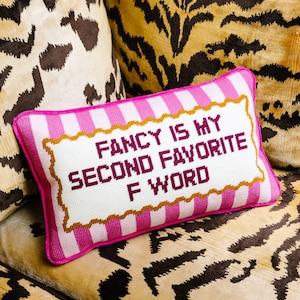May include: A rectangular decorative pillow with a white background and pink stripes. The pillow features the embroidered text "FANCY IS MY SECOND FAVORITE F WORD" in burgundy. The pillow is framed with a gold border and a pink edge.