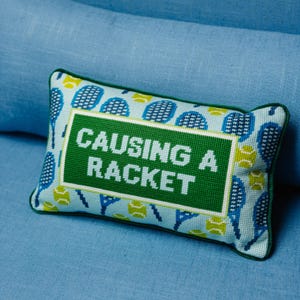 May include: A rectangular decorative pillow with a tennis theme. The pillow features a light blue background with tennis rackets and balls. The center has a green rectangle with the words "CAUSING A RACKET" in white.
