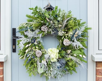 May include: A vibrant, circular wreath composed of various green, white, and blue foliage, including pinecones and hydrangea blooms. The wreath is displayed on a light blue door, creating a welcoming aesthetic. The door has a black handle.