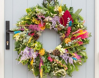 May include: A colorful, circular wreath with a variety of flowers, feathers, and pinecones hangs on a light gray door. The door is framed by white trim and flanked by windows. The exterior wall is made of red brick.