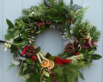 May include: A festive wreath composed of evergreen branches, pinecones, berries, and dried orange slices. The wreath is adorned with various green and red foliage, creating a natural and seasonal decoration. It hangs on a light blue door.