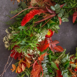 Puede incluir: Una corona de oto&ntilde;o con una variedad de vegetaci&oacute;n, hojas rojas y naranjas y flores secas. La corona est&aacute; hecha con eucalipto, bayas y otros elementos naturales.