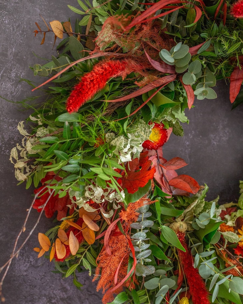 Puede incluir: Una corona de oto&ntilde;o con una variedad de vegetaci&oacute;n, hojas rojas y naranjas y flores secas. La corona est&aacute; hecha con eucalipto, bayas y otros elementos naturales.