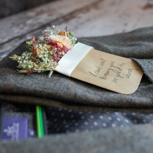 May include: A brown card with the text "I can't wait to marry you, see you at the church" is tucked into a gray suit jacket. A small bouquet of dried flowers is pinned to the card.