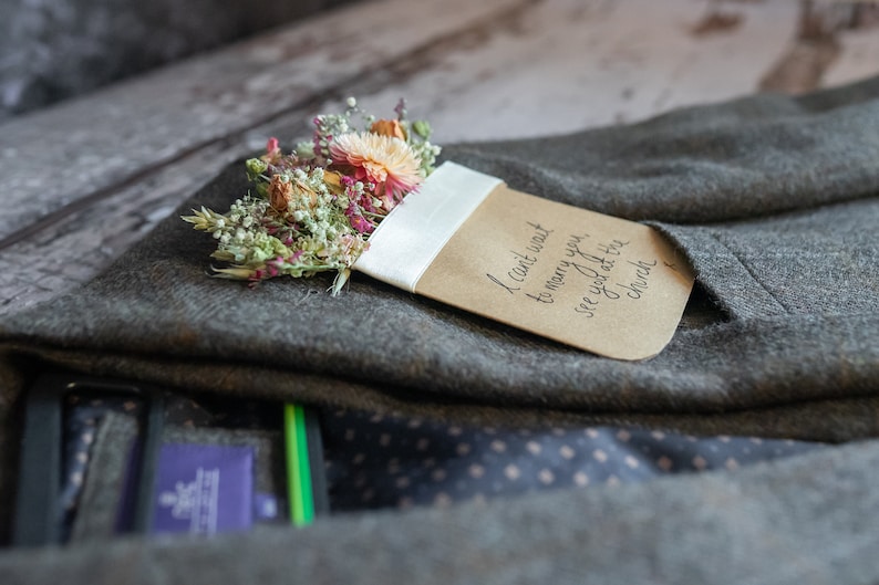 May include: A brown card with the text "I can't wait to marry you, see you at the church" is tucked into a gray suit jacket. A small bouquet of dried flowers is pinned to the card.