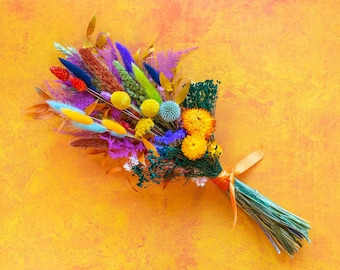 May include: A colorful bouquet of dried flowers in shades of yellow, blue, purple, and green. The flowers are tied together with a light orange ribbon.