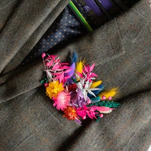 May include: A colorful floral boutonniere with pink, yellow, white, and blue flowers. The boutonniere is pinned to a brown tweed jacket.