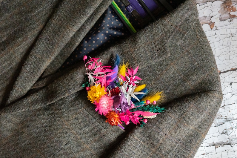 May include: A colorful floral boutonniere with pink, yellow, white, and blue flowers. The boutonniere is pinned to a brown tweed jacket.