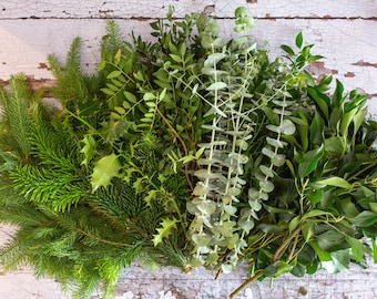 May include: An overhead view showcases a variety of fresh, green foliage. The selection includes evergreen branches, holly with spiky leaves, and eucalyptus with round leaves. The arrangement suggests a natural, seasonal theme, ideal for crafting or decoration.