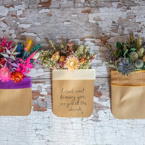 May include: Three small bouquets of dried flowers in brown paper bags with different colored ribbons. The middle bag has a handwritten message that reads "I can't wait to marry you, see you at the church x".