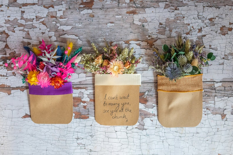 May include: Three small bouquets of dried flowers in brown paper bags with different colored ribbons. The middle bag has a handwritten message that reads "I can't wait to marry you, see you at the church x".