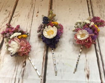 Puede incluir: Tres pinzas para el pelo plateadas con arreglos de flores secas. Cada pinza presenta una disposición diferente de flores rosas, moradas, blancas y amarillas.