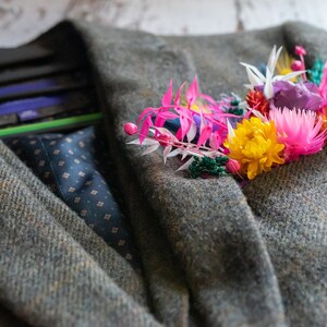 May include: A green tweed jacket with a colorful floral boutonniere. The boutonniere features pink, yellow, orange, white, and purple flowers.