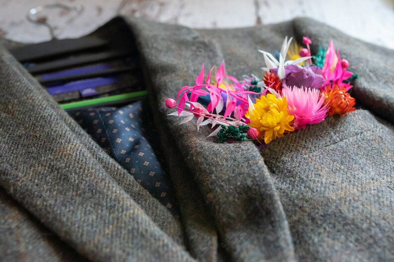 May include: A green tweed jacket with a colorful floral boutonniere. The boutonniere features pink, yellow, orange, white, and purple flowers.