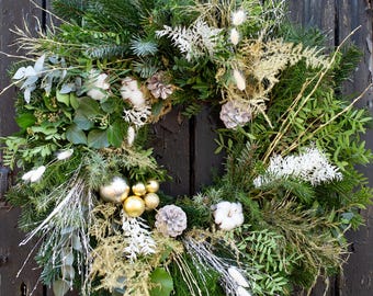 May include: A festive Christmas wreath with a variety of greenery, including pine, ivy, and sprigs of white and gold accents. The wreath is adorned with gold and silver ornaments, pinecones, and cotton-like accents. The wreath is displayed on a dark wooden door.