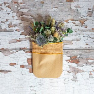 May include: A bouquet of dried flowers, including purple thistle, green eucalyptus, and wheat, arranged in a brown paper bag with a yellow burlap trim.