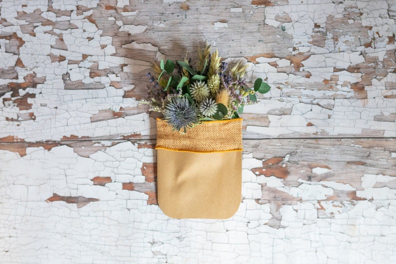 May include: A bouquet of dried flowers, including purple thistle, green eucalyptus, and wheat, arranged in a brown paper bag with a yellow burlap trim.