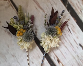 Puede incluir: Dos pinzas para el pelo plateadas con arreglos de flores secas. Los arreglos presentan flores blancas, amarillas, azules y moradas.