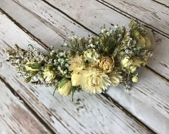 May include: A crescent-shaped floral crown made with dried flowers in shades of cream and green. The crown is made with small white flowers, larger cream-colored flowers, and green foliage.
