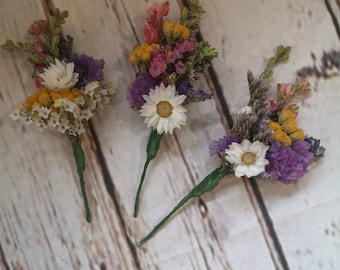 Puede incluir: Tres pequeños ramos de flores secas con margaritas blancas, flores moradas, rosas y amarillas. Los ramos están atados con una cinta verde y tienen tallos largos.