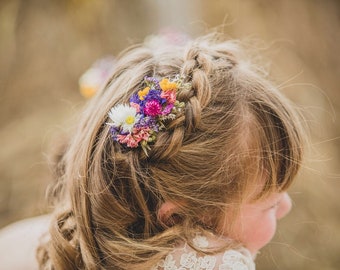 Puede incluir: Una niña con cabello rubio que lleva un vestido blanco con una trenza floral en el cabello. La trenza está decorada con pequeñas flores moradas, rosas, blancas y amarillas.