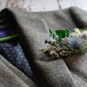 May include: A green tweed jacket with a small bouquet of dried flowers pinned to the lapel. The flowers are a mix of purple, white, and green.