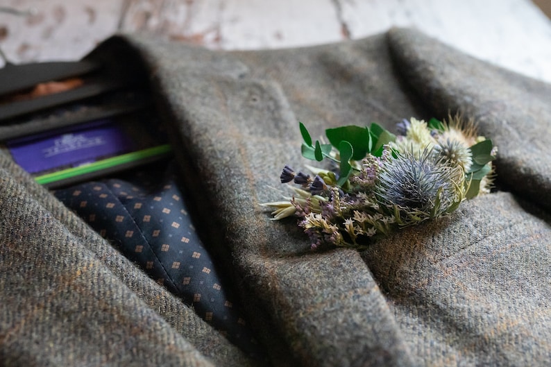 May include: A green tweed jacket with a small bouquet of dried flowers pinned to the lapel. The flowers are a mix of purple, white, and green.