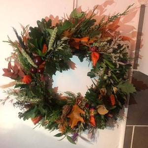 Puede incluir: Una corona de oto&ntilde;o hecha con elementos naturales, incluyendo hojas verdes, ramas marrones, bayas naranjas y rojas y flores secas.