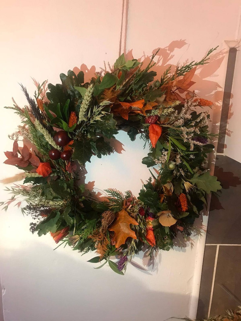 Puede incluir: Una corona de oto&ntilde;o hecha con elementos naturales, incluyendo hojas verdes, ramas marrones, bayas naranjas y rojas y flores secas.