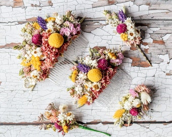 Puede incluir: Cuatro peines para el cabello decorados con flores secas. Los peines son de plástico y tienen una base blanca. Las flores son una mezcla de morado, amarillo y rosa.