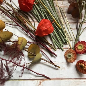 Puede incluir: Flores y follaje secos en tonos rojos, naranjas y amarillos, incluyendo hojas de eucalipto, bayas rojas y peque&ntilde;as flores naranjas.