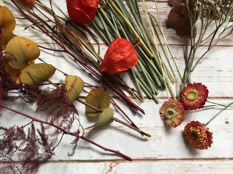 Puede incluir: Flores y follaje secos en tonos rojos, naranjas y amarillos, incluyendo hojas de eucalipto, bayas rojas y peque&ntilde;as flores naranjas.
