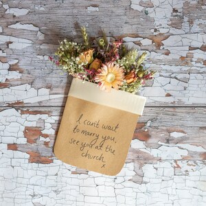 May include: A brown paper envelope with a white ribbon tied around it. The envelope contains a bouquet of dried flowers and a handwritten message that reads "I can't wait to marry you, see you at the church x".