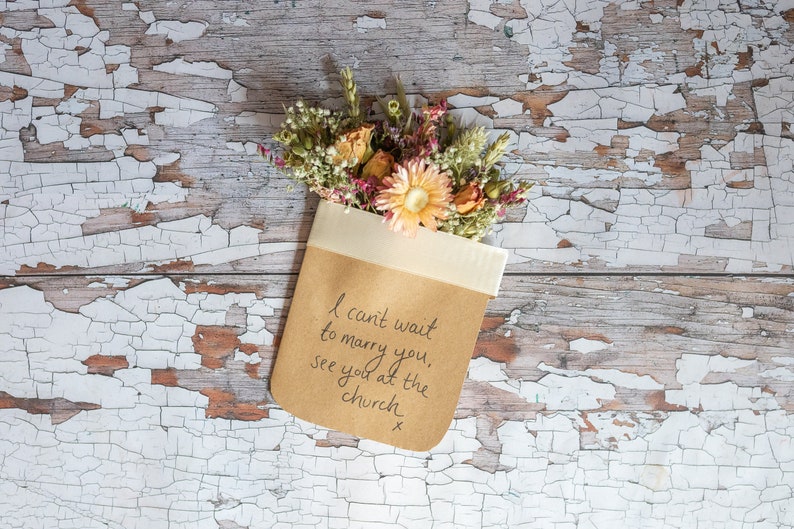 May include: A brown paper envelope with a white ribbon tied around it. The envelope contains a bouquet of dried flowers and a handwritten message that reads "I can't wait to marry you, see you at the church x".