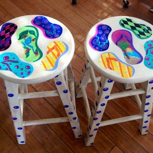 May include: Two wooden stools painted with blue polka dots and decorated with colorful flip-flop designs. The flip-flops are painted with various patterns, including a tropical scene, a green frog, and a swirl design.