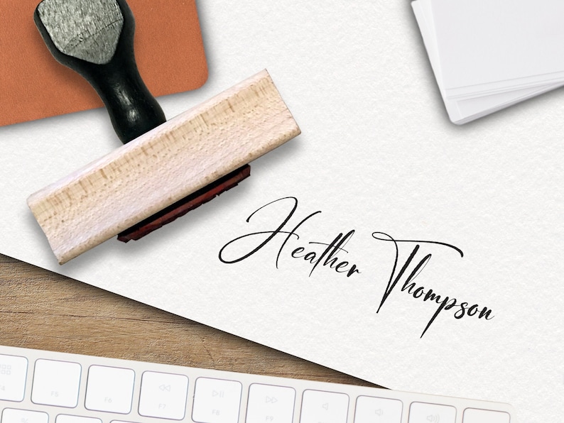 Op de afbeelding: Een houten stempel met een zwarte handgreep en een rood inktkussen. De stempel wordt gebruikt om een ​​handtekening te maken die "Heather Thompson" luidt.