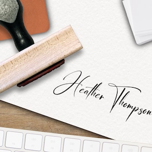 Op de afbeelding: Een houten zelf-inktende stempel met een zwarte handgreep en een rood inktkussen. De stempel wordt gebruikt om een ​​handtekening te maken die "Heather Thompson" luidt.