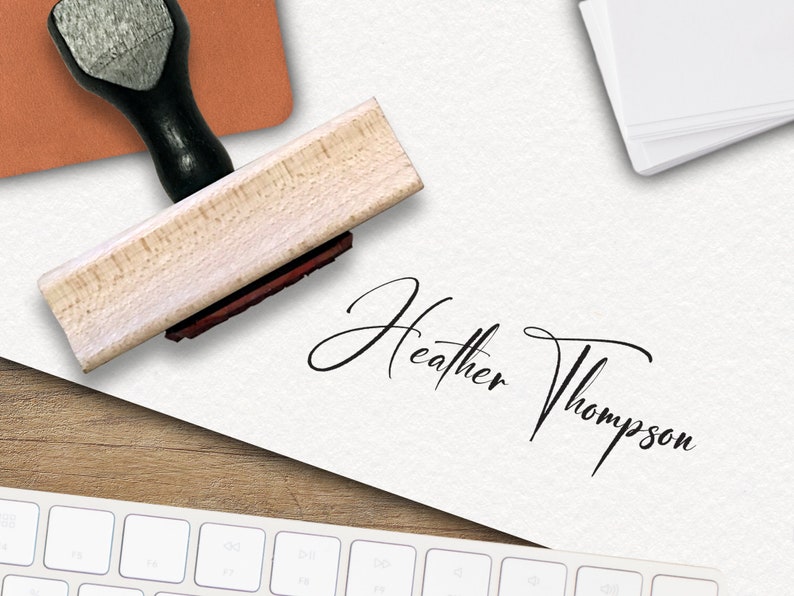 Op de afbeelding: Een houten zelf-inktende stempel met een zwarte handgreep en een rood inktkussen. De stempel wordt gebruikt om een ​​handtekening te maken die "Heather Thompson" luidt.