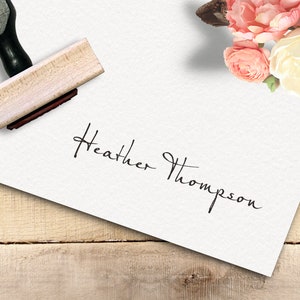 Op de afbeelding: Een witte kaart met de handgeschreven handtekening "Heather Thompson" erop. Een houten stempel met een zwarte handgreep ligt op de kaart. De kaart ligt op een houten oppervlak met roze en witte bloemen op de achtergrond.