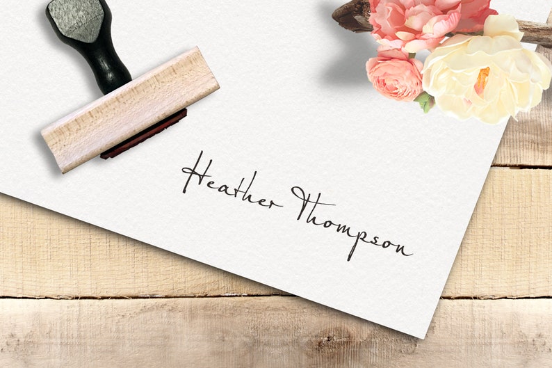 Op de afbeelding: Een witte kaart met de handgeschreven handtekening "Heather Thompson" erop. Een houten stempel met een zwarte handgreep ligt op de kaart. De kaart ligt op een houten oppervlak met roze en witte bloemen op de achtergrond.