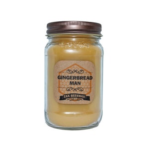 May include: A clear glass jar candle with a bronze lid. The candle is filled with yellow beeswax and has a label that reads "GINGERBREAD MAN" and "ZAX BEESWAX". The candle is a natural beeswax candle.
